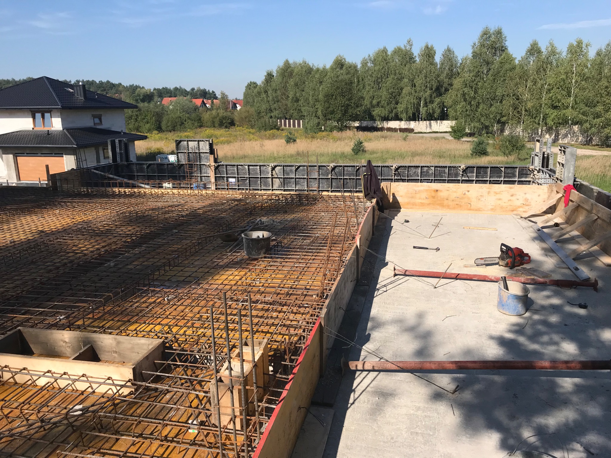 Zbrojenie fundamentu przygotowane do zalania betonem, widoczne szalunki, pręty zbrojeniowe, narzędzia budowlane i w oddali dom w budowie.