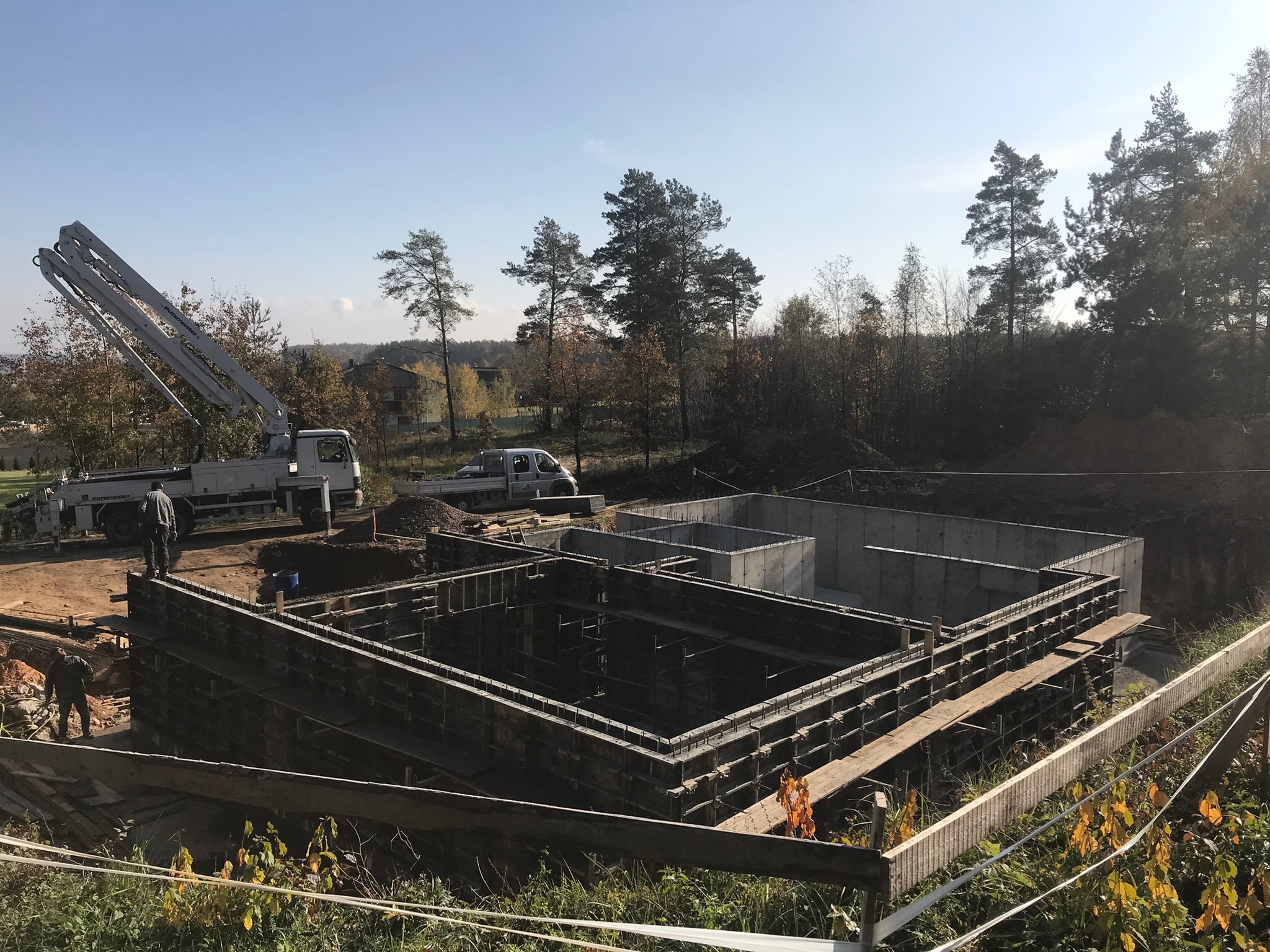 Wykop pod fundamenty domu z szalunkiem, widoczny betonowy wylew oraz pompa do betonu na tle drzew.