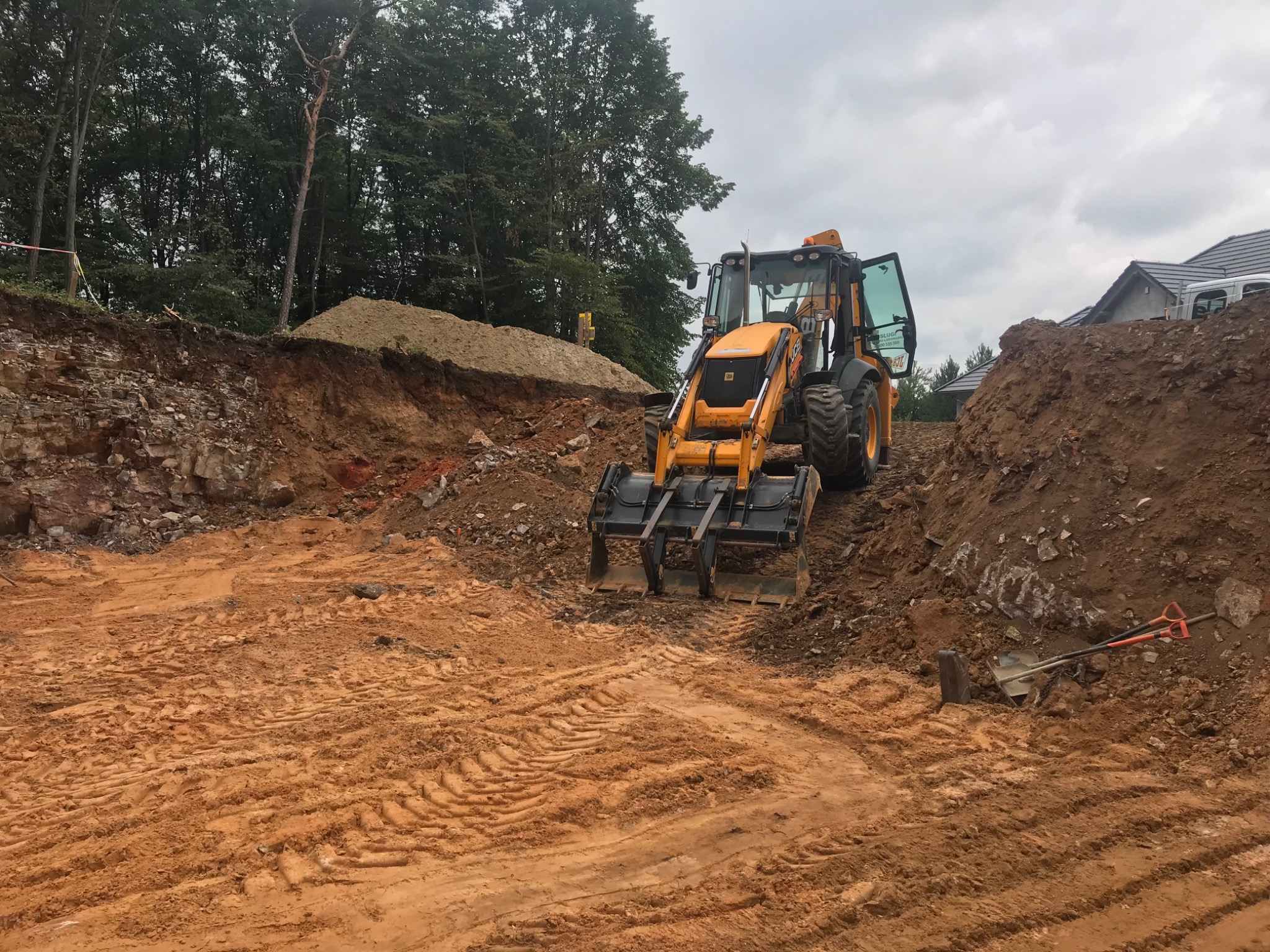 Żółta koparko-ładowarka JCB na placu budowy, przygotowanie terenu pod fundamenty, widoczne ślady opon w błotnistym podłożu, w tle drzewa i częściowo zarys domu.