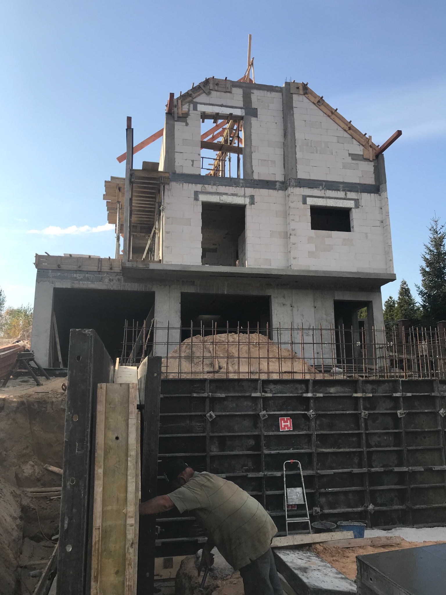 Wznoszenie dwukondygnacyjnego domu z betonu komórkowego, widoczne szalunki fundamentowe, pracownik przy szalunkach, częściowo ukończone ściany parteru i piętra, konstrukcja dachu w trakcie budowy.