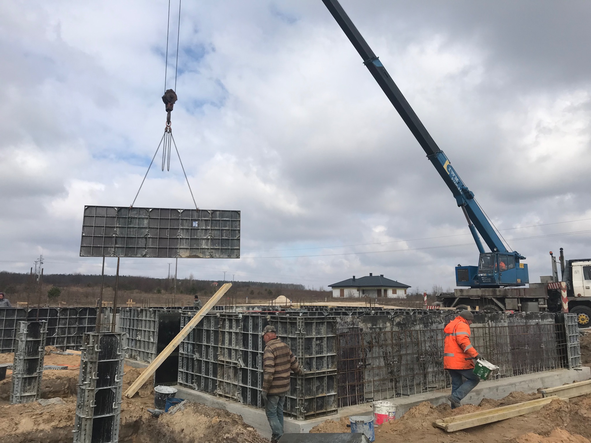 Montaż szalunków fundamentowych za pomocą dźwigu na placu budowy, w tle dom jednorodzinny.