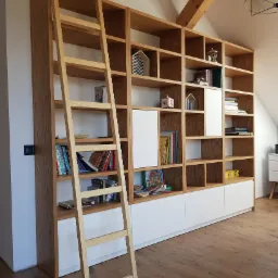 Duża, drewniana biblioteczka z drabinką w jasnym odcieniu drewna, z białymi, dolnymi szafkami, umieszczona w pokoju na poddaszu z drewnianą więźbą dachową.