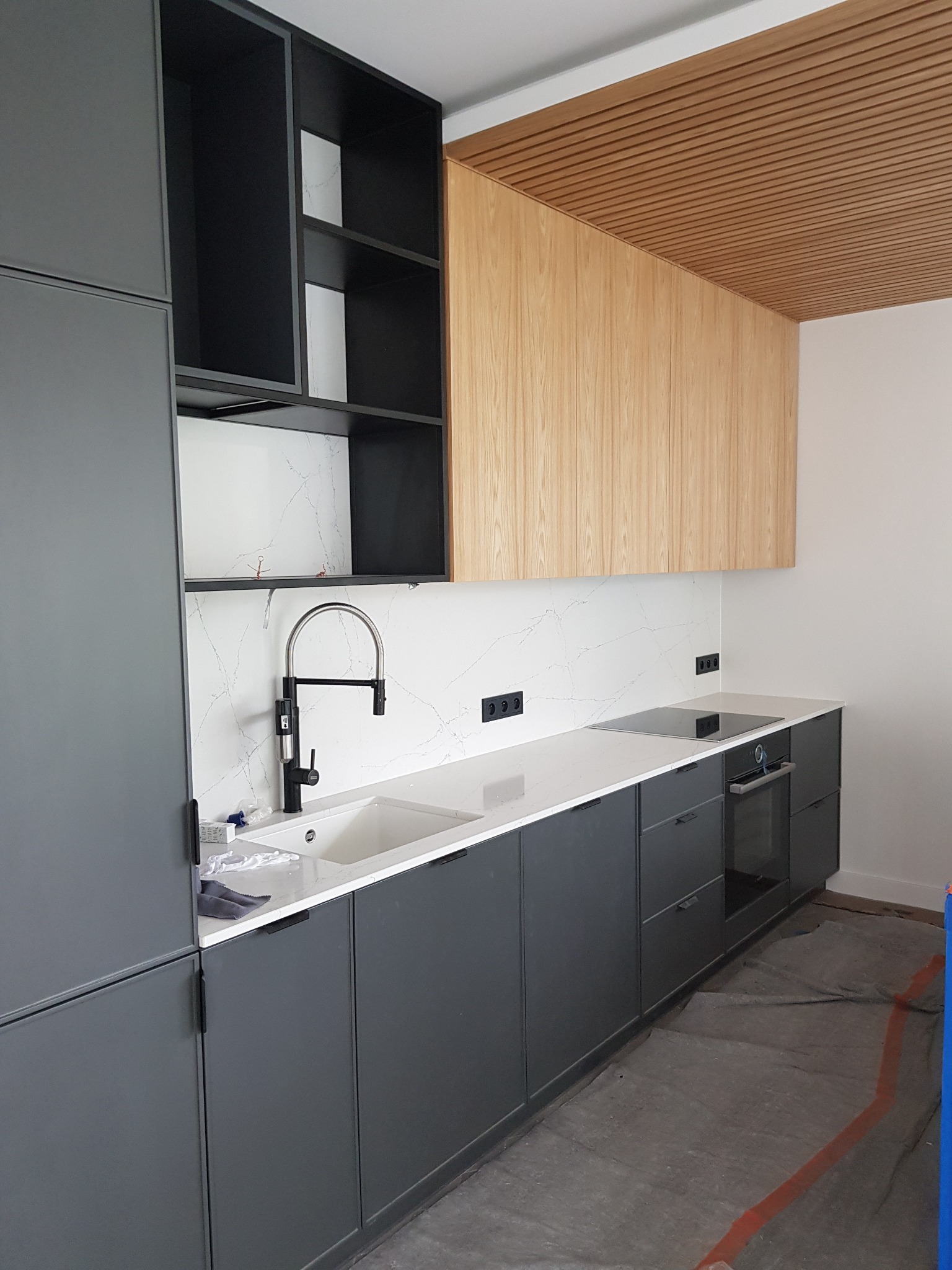 Nowoczesna kuchnia z szarymi szafkami dolnymi i czarnymi otwartymi półkami górnymi, blatem z białego marmuru i drewnianymi szafkami górnymi, w trakcie montażu, z widocznym zabezpieczeniem podłogi.