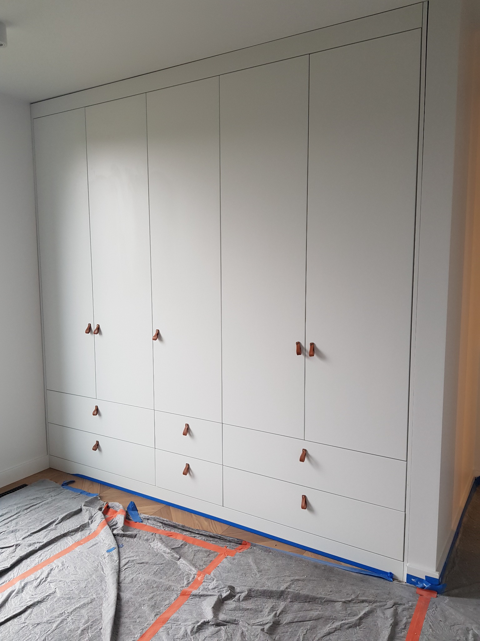 Biała zabudowa szafy z szufladami, z minimalistycznymi skórzanymi uchwytami, zabezpieczona folią malarską i taśmą na podłodze podczas prac wykończeniowych.