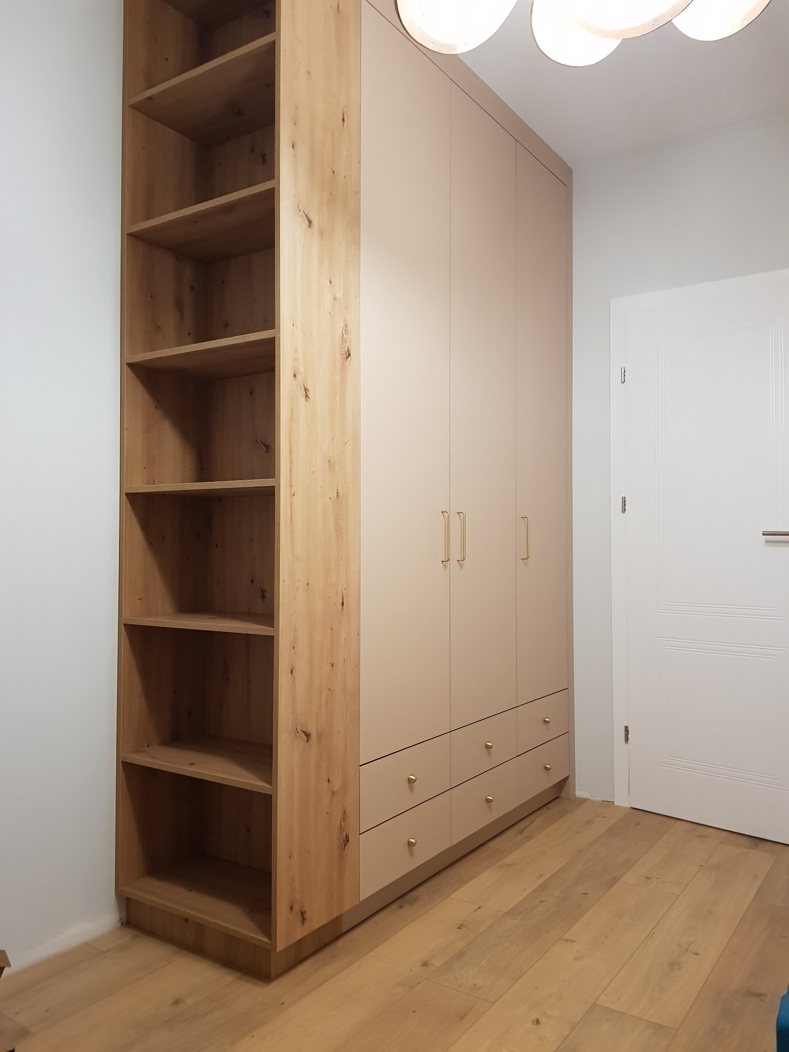 Beżowa zabudowa ścienna z szafami i otwartym regałem z drewna o widocznej strukturze usłojenia, złote uchwyty, drewniana podłoga, białe ściany i drzwi.