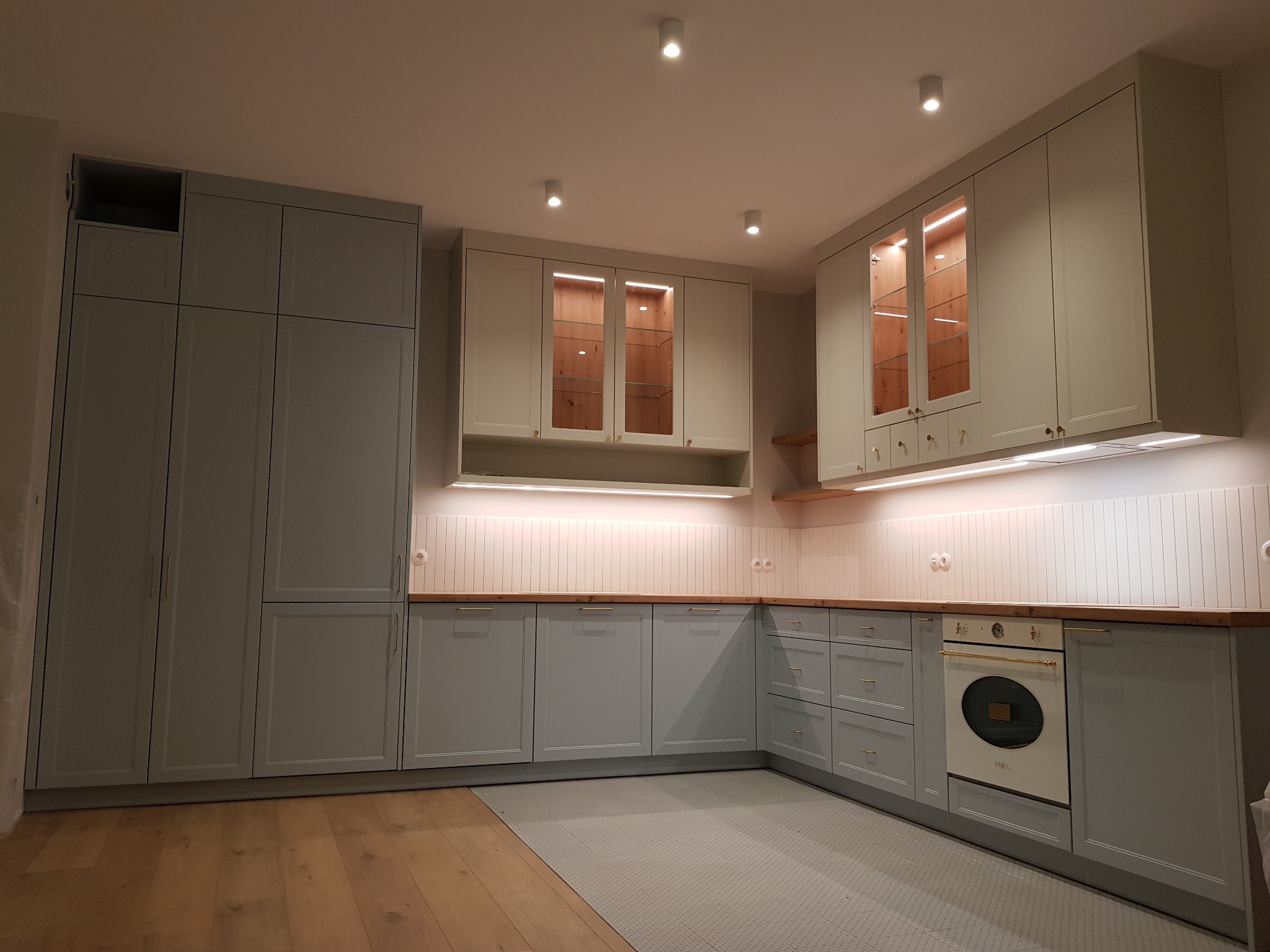 Kompletna zabudowa kuchenna w odcieniach szarości z drewnianymi blatami i oświetleniem podszafkowym, z widocznymi szklanymi witrynami górnych szafek podświetlonymi od wewnątrz.