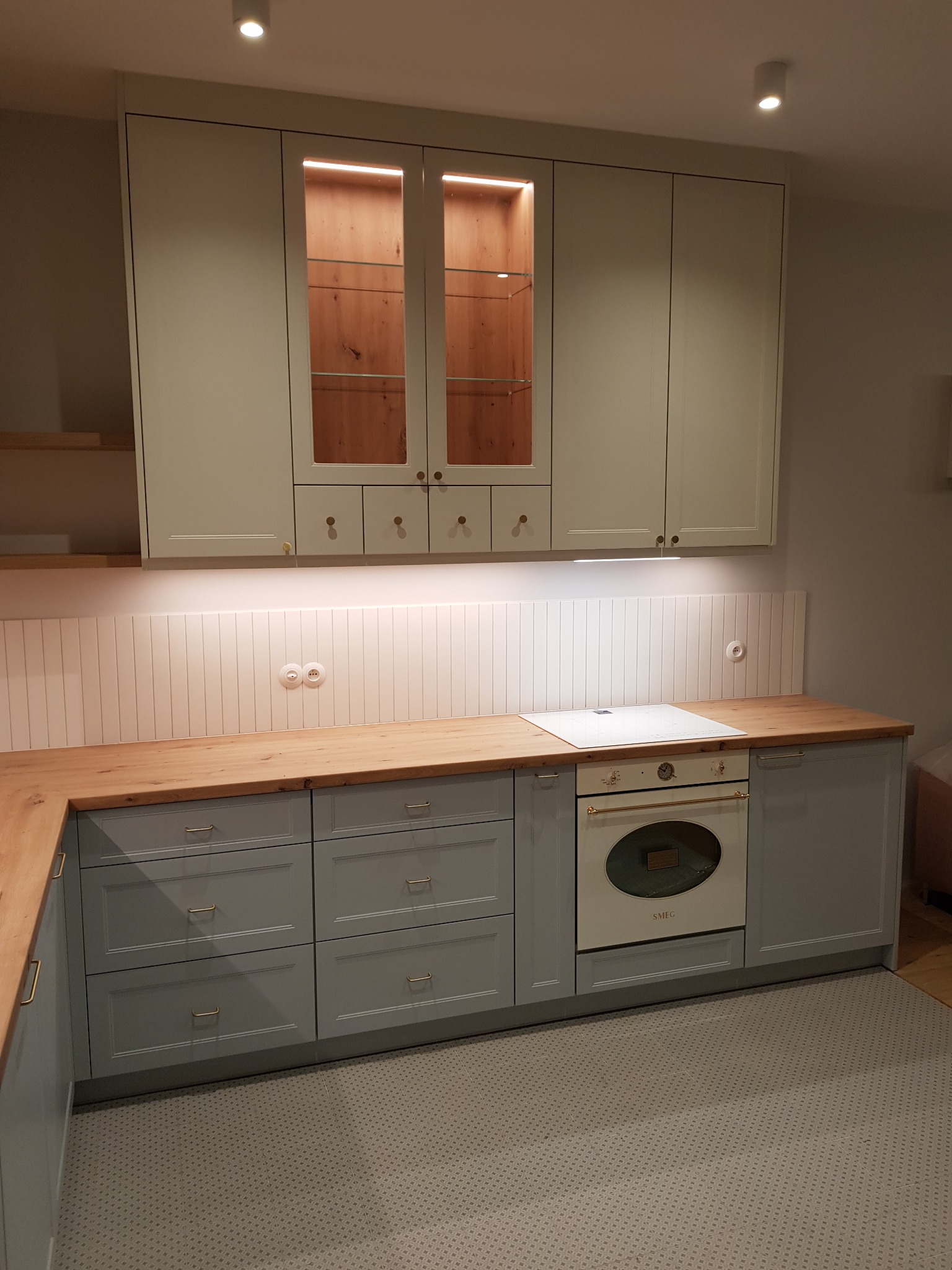 Kuchnia w odcieniach szarości z drewnianym blatem i oświetleniem podszafkowym, przeszklona szafka górna z podświetleniem, styl retro.