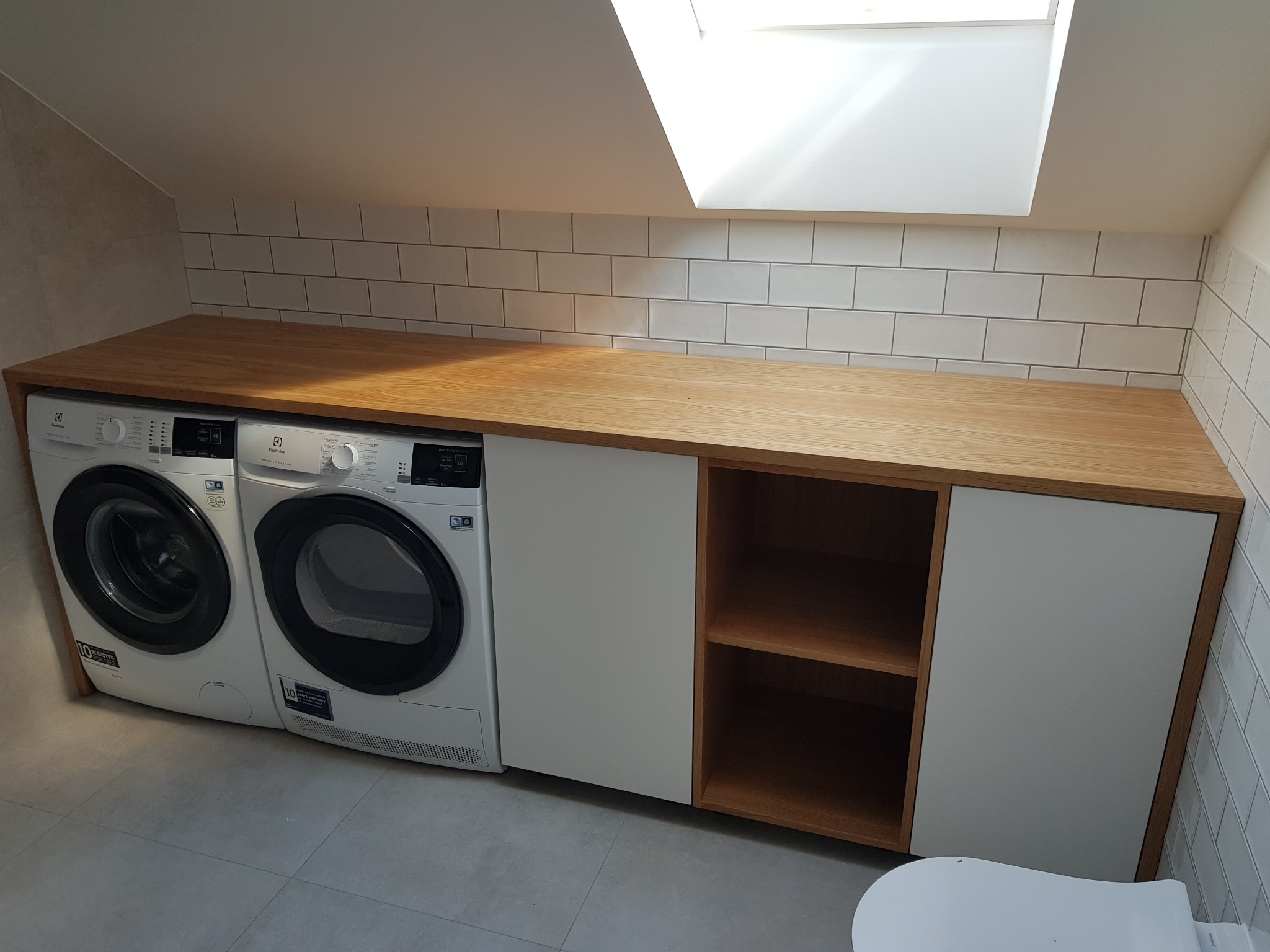 Pralka i suszarka zabudowane pod drewnianym blatem w łazience na poddaszu, widoczne białe płytki na ścianie i okno dachowe.