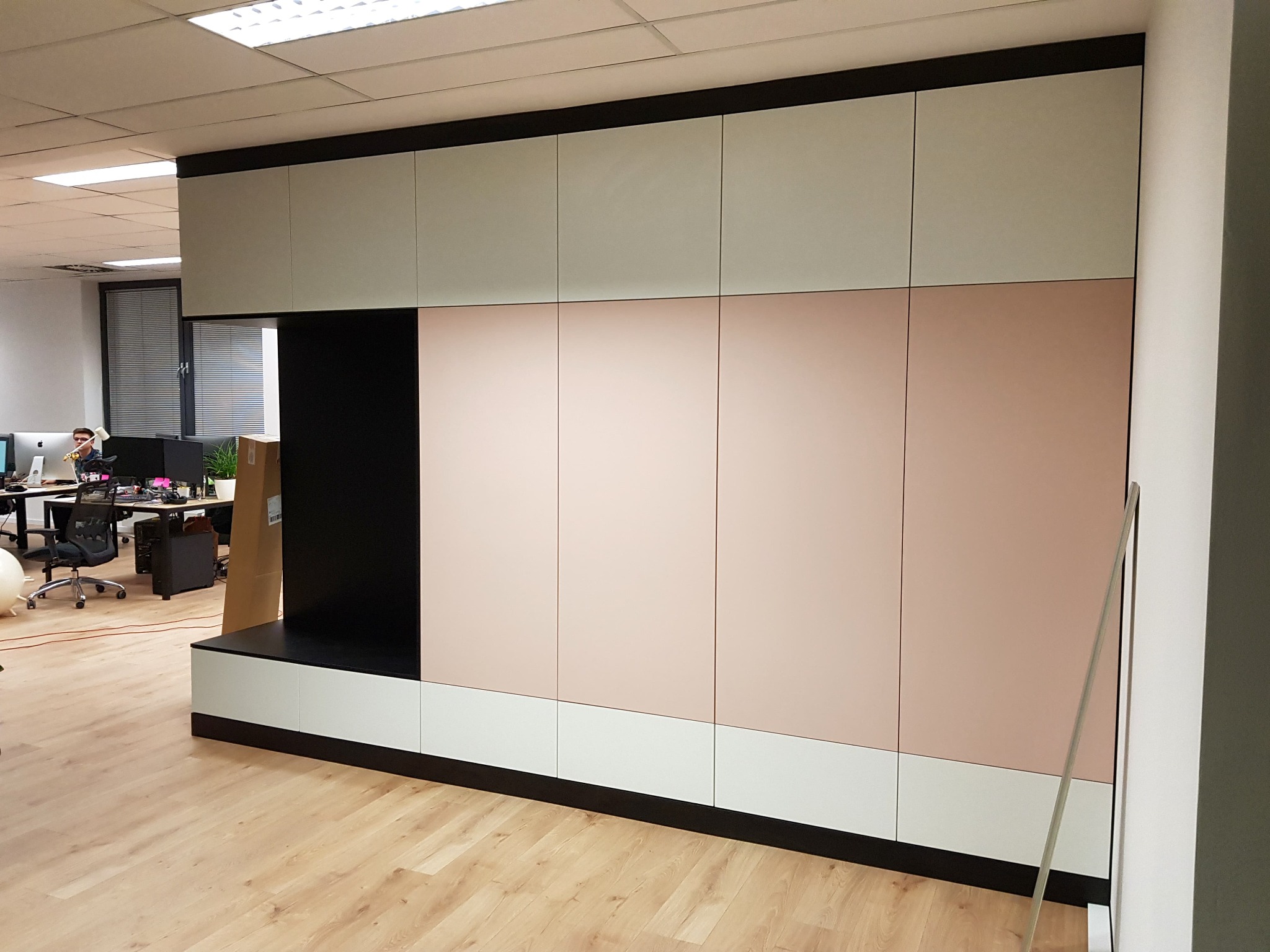 Wielobarwna zabudowa meblowa w biurze: pastelowe różowe i szare fronty szaf, czarne elementy konstrukcyjne, jasna podłoga drewnopodobna, widok na stanowiska pracy w tle.