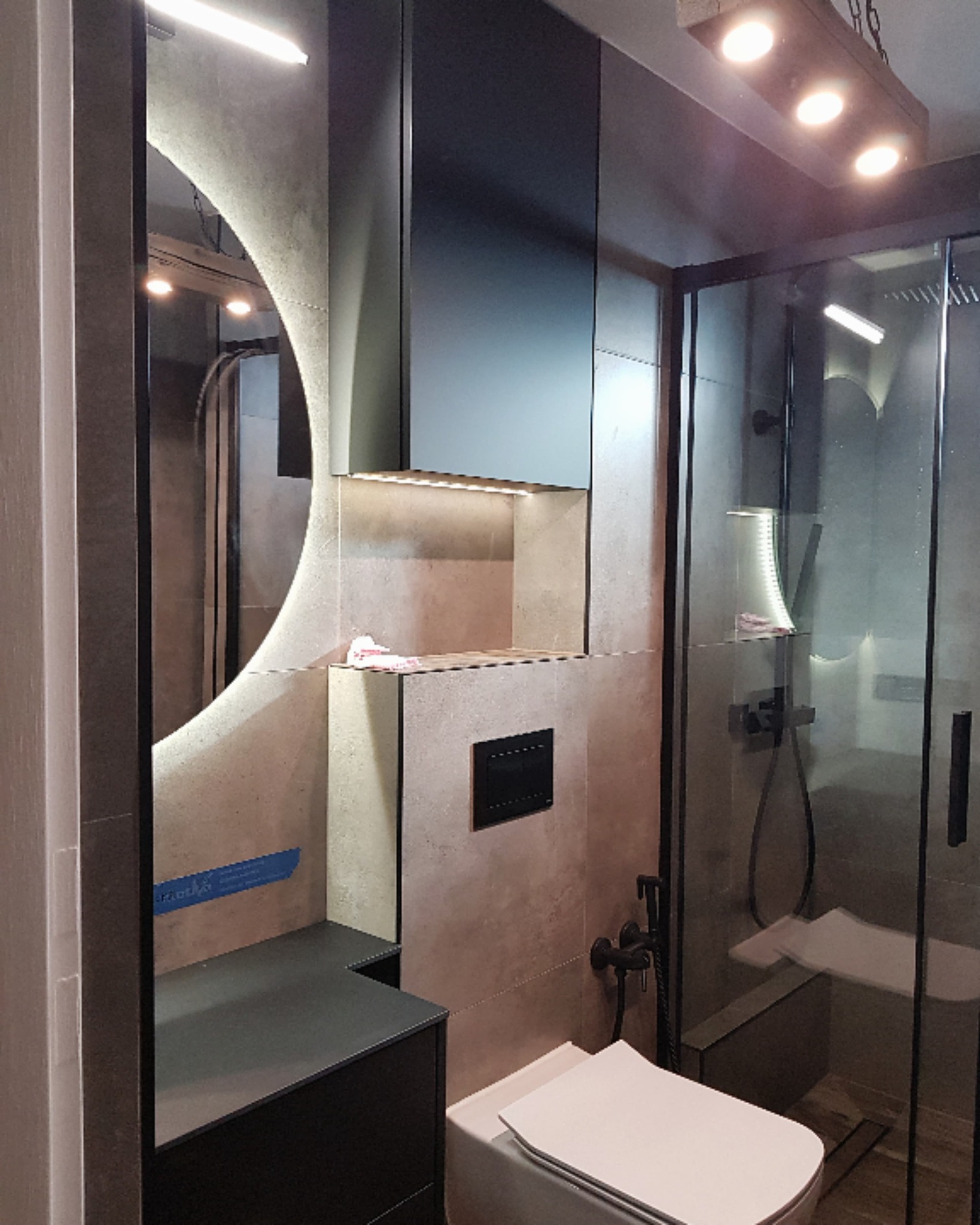 Nowoczesna łazienka z podświetlanym lustrem o zaokrąglonym kształcie, czarnymi szafkami pod umywalką, kabiną prysznicową z czarnymi ramami i toaletą. Na ścianie widoczna czarna armatura i beżowe...