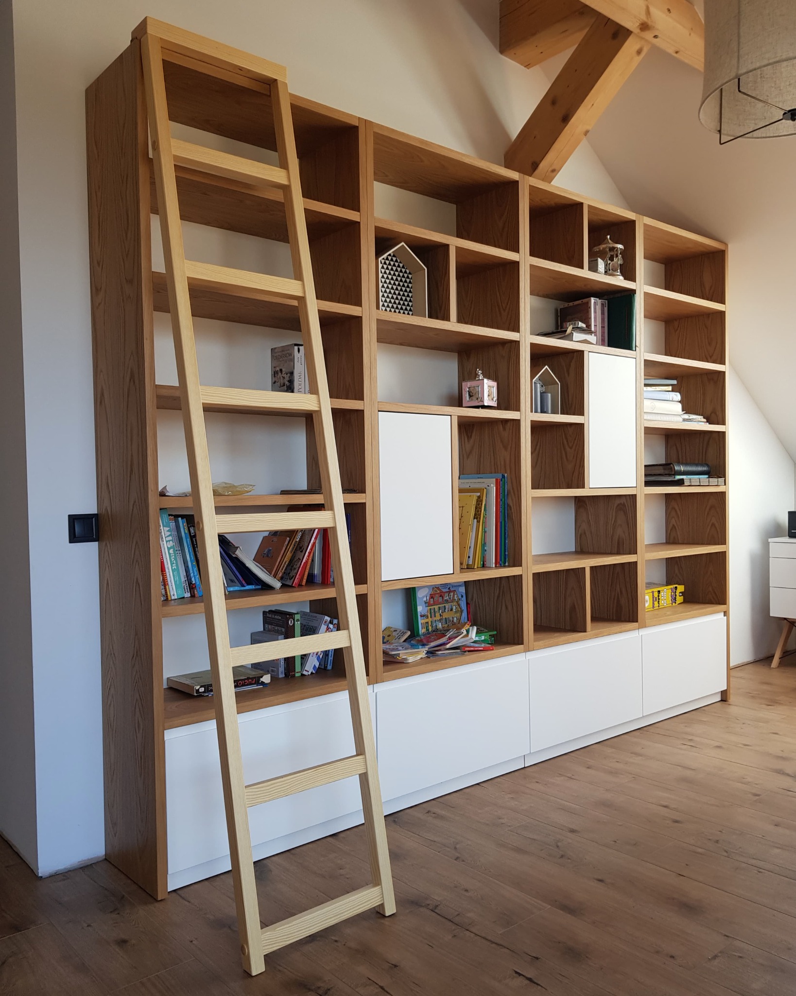 Duża, drewniana biblioteczka z drabinką w jasnym odcieniu drewna, z białymi, dolnymi szafkami, umieszczona w pokoju na poddaszu z drewnianą więźbą dachową.