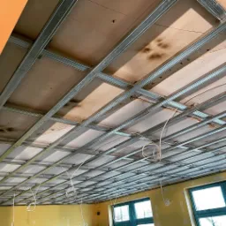 Metalowa konstrukcja sufitu podwieszanego z widocznymi płytami, częściowo zamontowanymi, z przewodami elektrycznymi zwisającymi w pomieszczeniu z żółtymi ścianami i ciemnozielonymi ramami okiennymi.