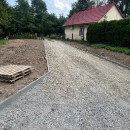 Przygotowany podjazd z utwardzoną nawierzchnią z kruszywa, obrzeża betonowe, w tle dom z czerwonym dachem i zielone drzewa.
