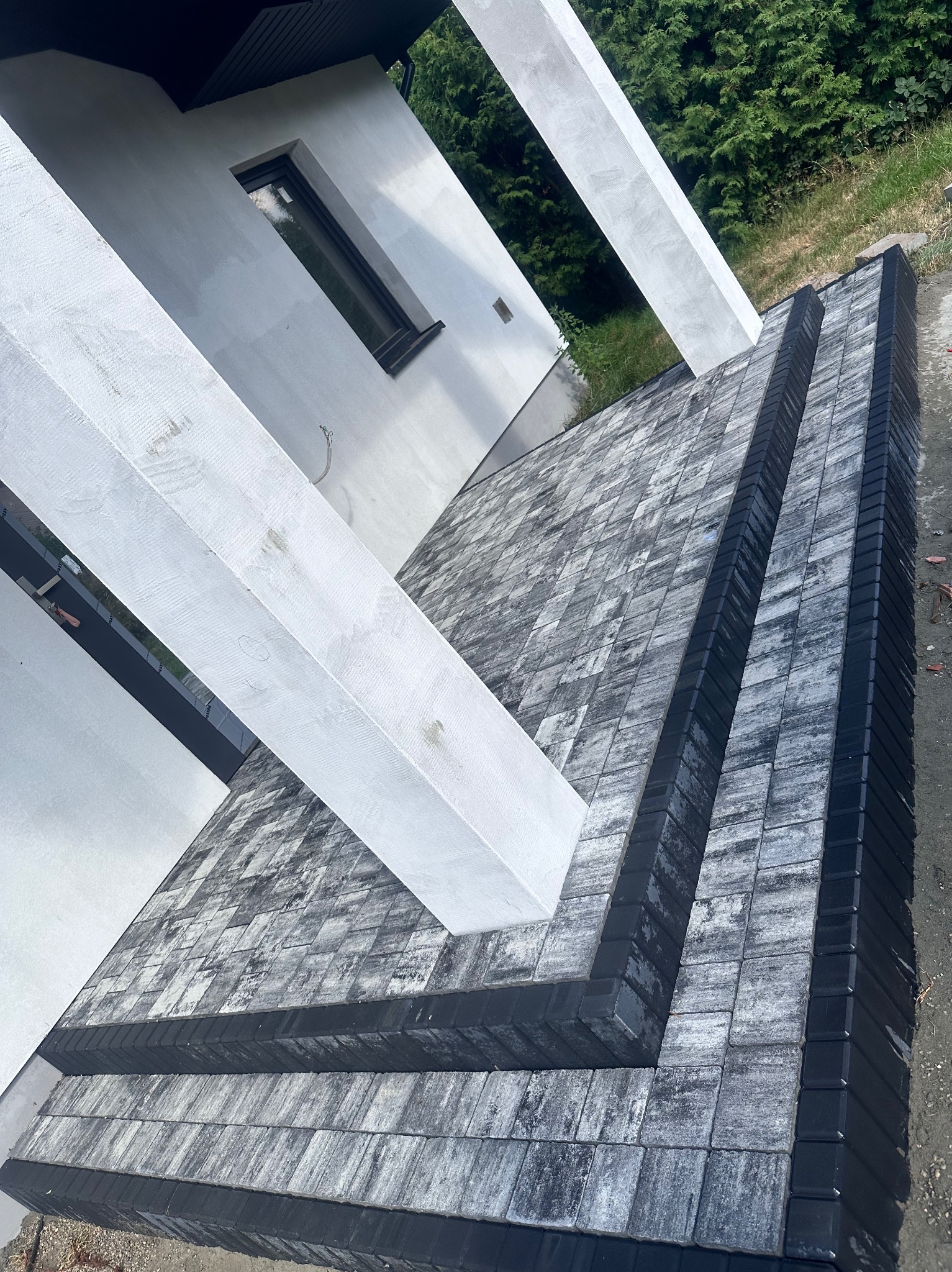 Ułożona kostka brukowa w odcieniach szarości z czarnym obrzeżem, tworząca schody przed nowoczesnym białym budynkiem z betonowymi filarami.