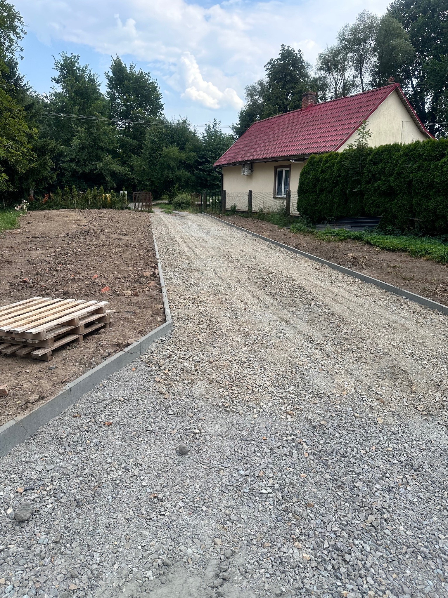 Przygotowany podjazd z utwardzoną nawierzchnią z kruszywa, obrzeża betonowe, w tle dom z czerwonym dachem i zielone drzewa.