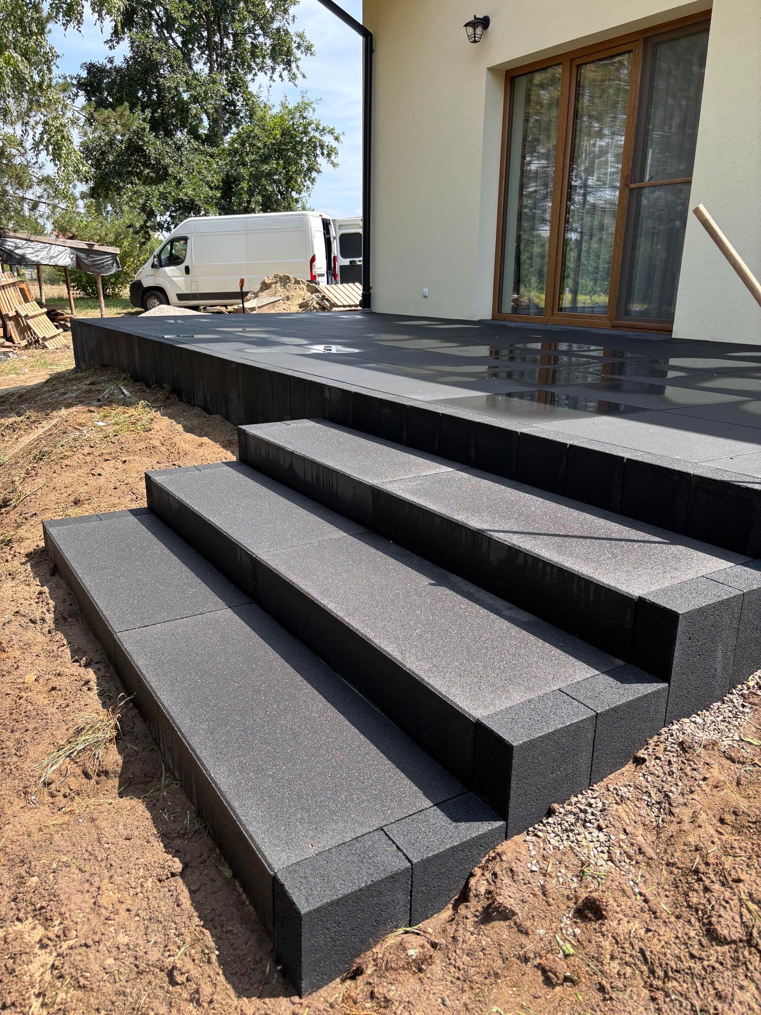 Nowoczesne, grafitowe schody z kostki brukowej prowadzące do domu z dużym oknem tarasowym, widoczny fragment elewacji i biały bus w tle.