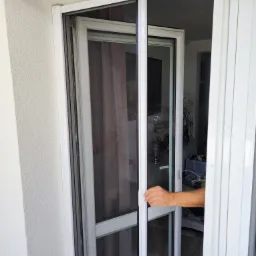 Biała, rozsuwana moskitiera okienna zamontowana na balkonie, widok z bliska na mechanizm przesuwny i dłoń osoby obsługującej system.