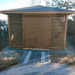 Drewniana altana z brązową roletą zewnętrzną w pozycji zamkniętej, widoczna z poziomu ścieżki z kostki brukowej, z dużym kamieniem po lewej stronie.