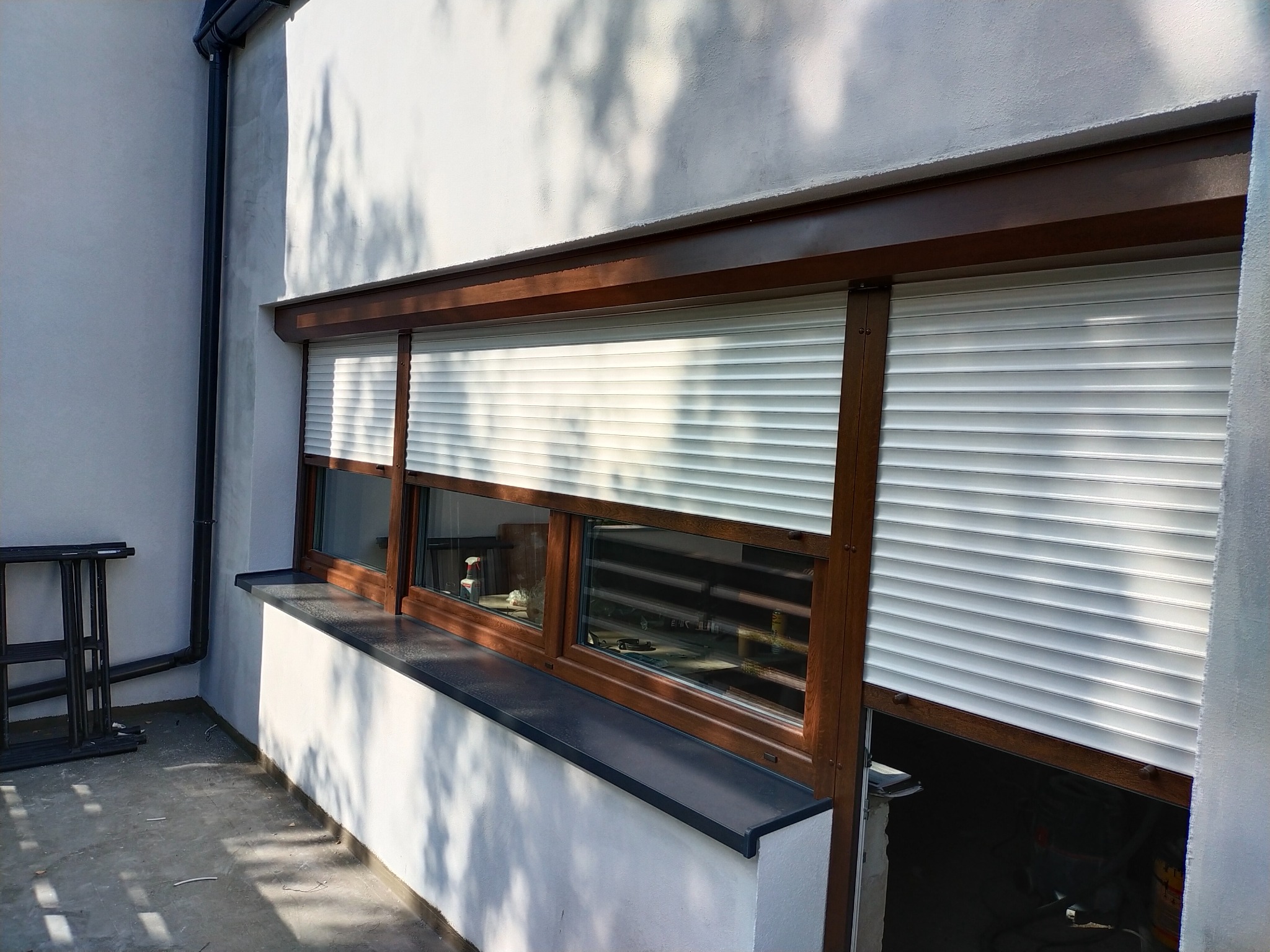 Białe rolety zewnętrzne z brązowymi ramami okiennymi, częściowo opuszczone, widoczne z zewnątrz budynku o jasnej elewacji w słoneczny dzień.