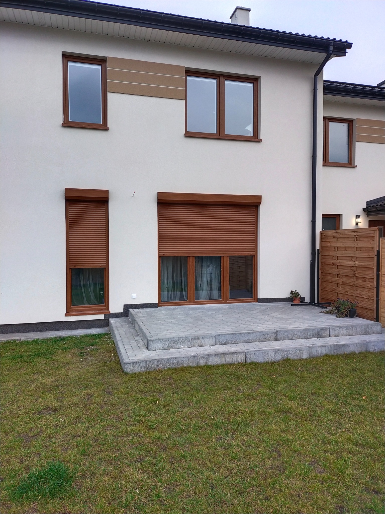 Elewacja domu z częściowo opuszczonymi brązowymi roletami zewnętrznymi, widoczny fragment tarasu wyłożonego szarą kostką brukową i trawnik.