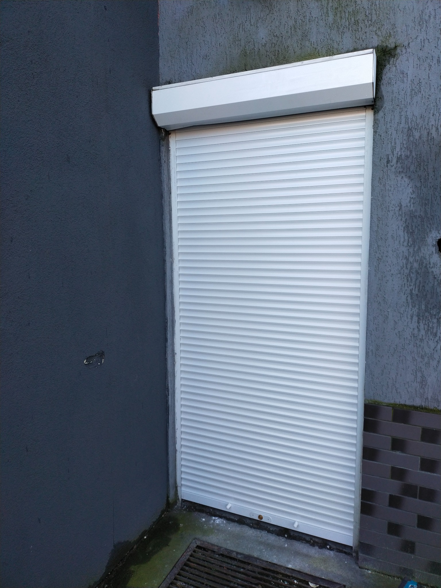 Biała roleta częściowo opuszczona na tle szarej ściany z widocznymi śladami wilgoci i kratką ściekową u dołu.