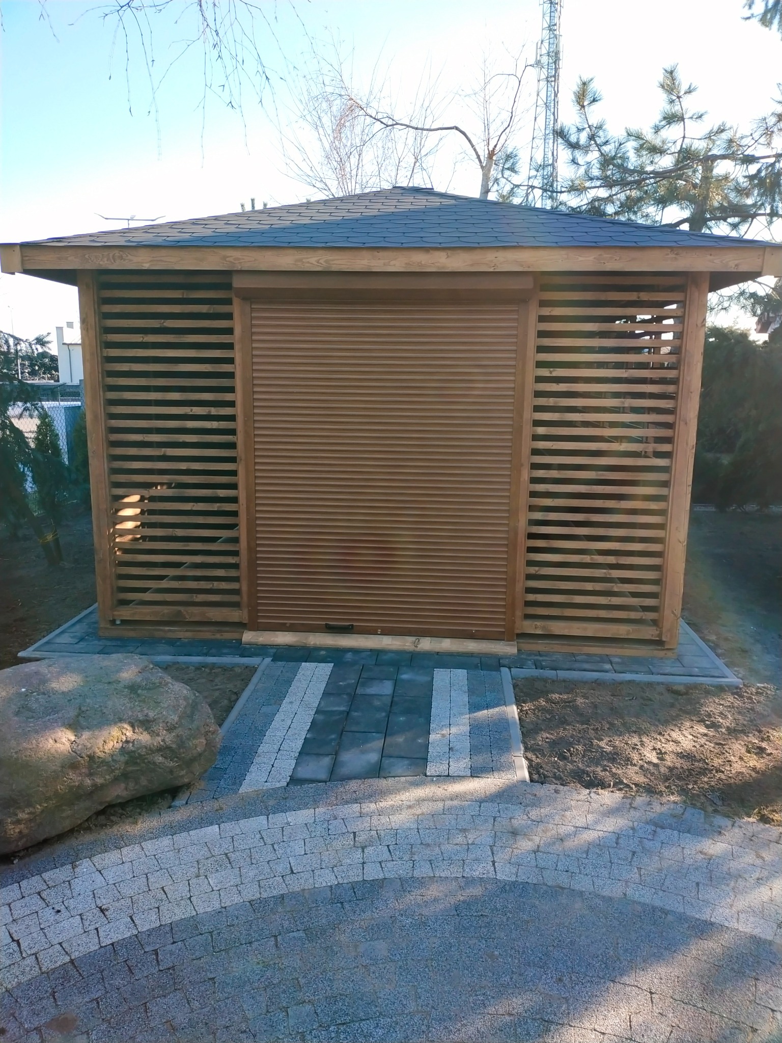 Drewniana altana z brązową roletą zewnętrzną w pozycji zamkniętej, widoczna z poziomu ścieżki z kostki brukowej, z dużym kamieniem po lewej stronie.