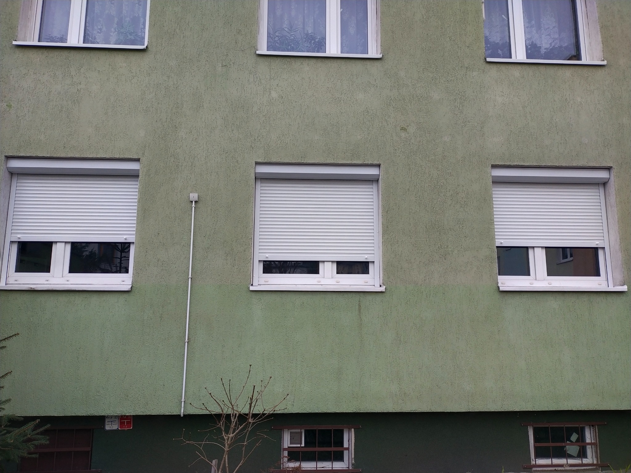 Elewacja budynku z trzema oknami na parterze i trzema na pierwszym piętrze, okna na pierwszym piętrze z częściowo opuszczonymi białymi roletami.