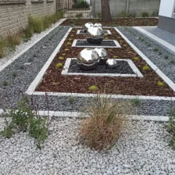 Nowoczesny ogród z geometrycznymi rabatami wypełnionymi korą, czarnym grysem i srebrnymi kulami, otoczony białym i szarym kamieniem.