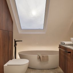 Usługi Projektowe - Nowoczesna łazienka na poddaszu z oknem dachowym, wolnostojącą wanną i podwieszaną toaletą. Umywalka z drewnianą szafką i kamienny dywanik.