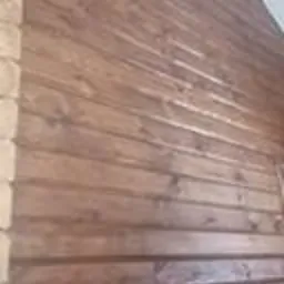 Fragment ściany zewnętrznej domu z bali, widoczne połączenia bali, pokryte brązowym impregnatem zabezpieczającym drewno.