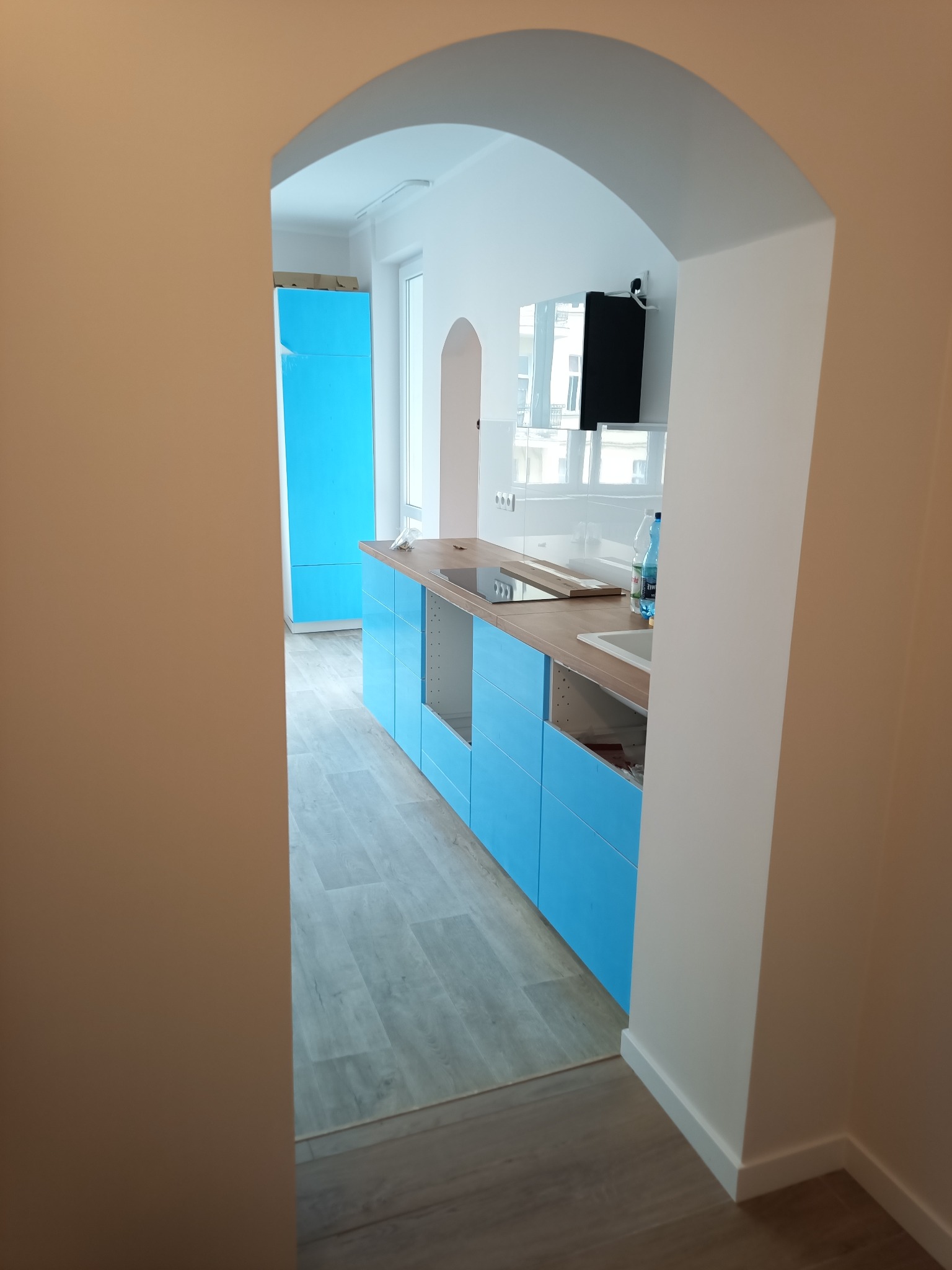 Widok na kuchnię z niebieskimi szafkami, drewnianym blatem, przez łukowe przejście w ścianie, widoczne elementy wykończenia wnętrz.