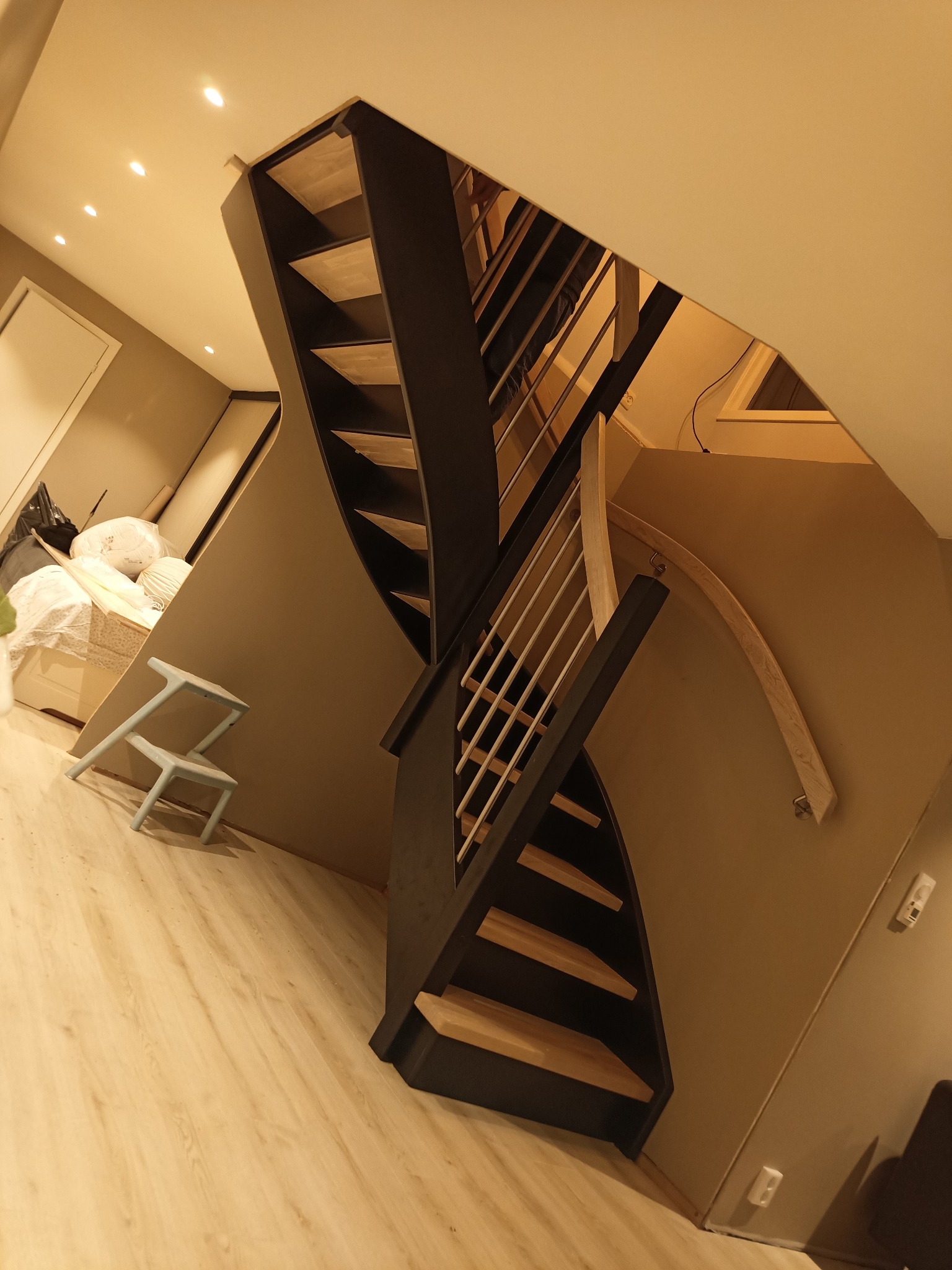 Nowoczesne, dwukierunkowe schody z ciemnymi bokami i jasnymi drewnianymi stopniami, w tle widoczna sypialnia i niebieski stołek.