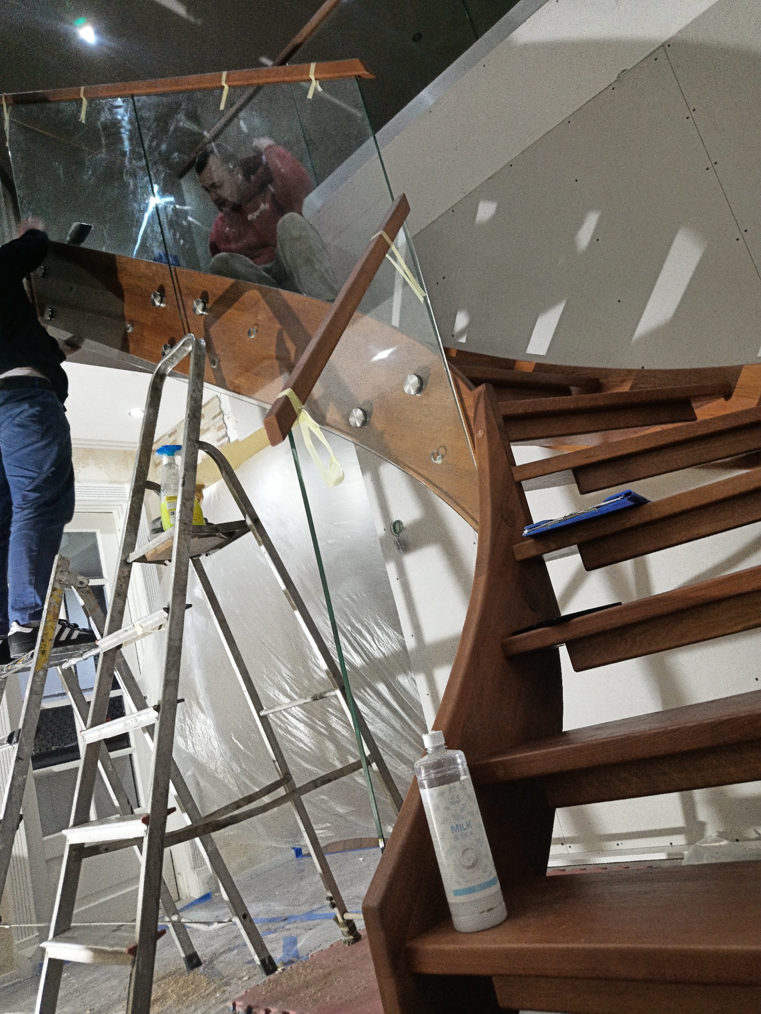 Montaż szklanej balustrady na drewnianych schodach kręconych, widoczna drabina, narzędzia i pracujący monter