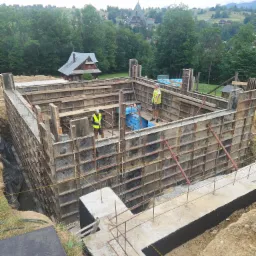 Budowa fundamentów: Dwóch pracowników w kamizelkach odblaskowych wewnątrz szalunku fundamentowego z widokiem na górski krajobraz i drewniany dom w tle.