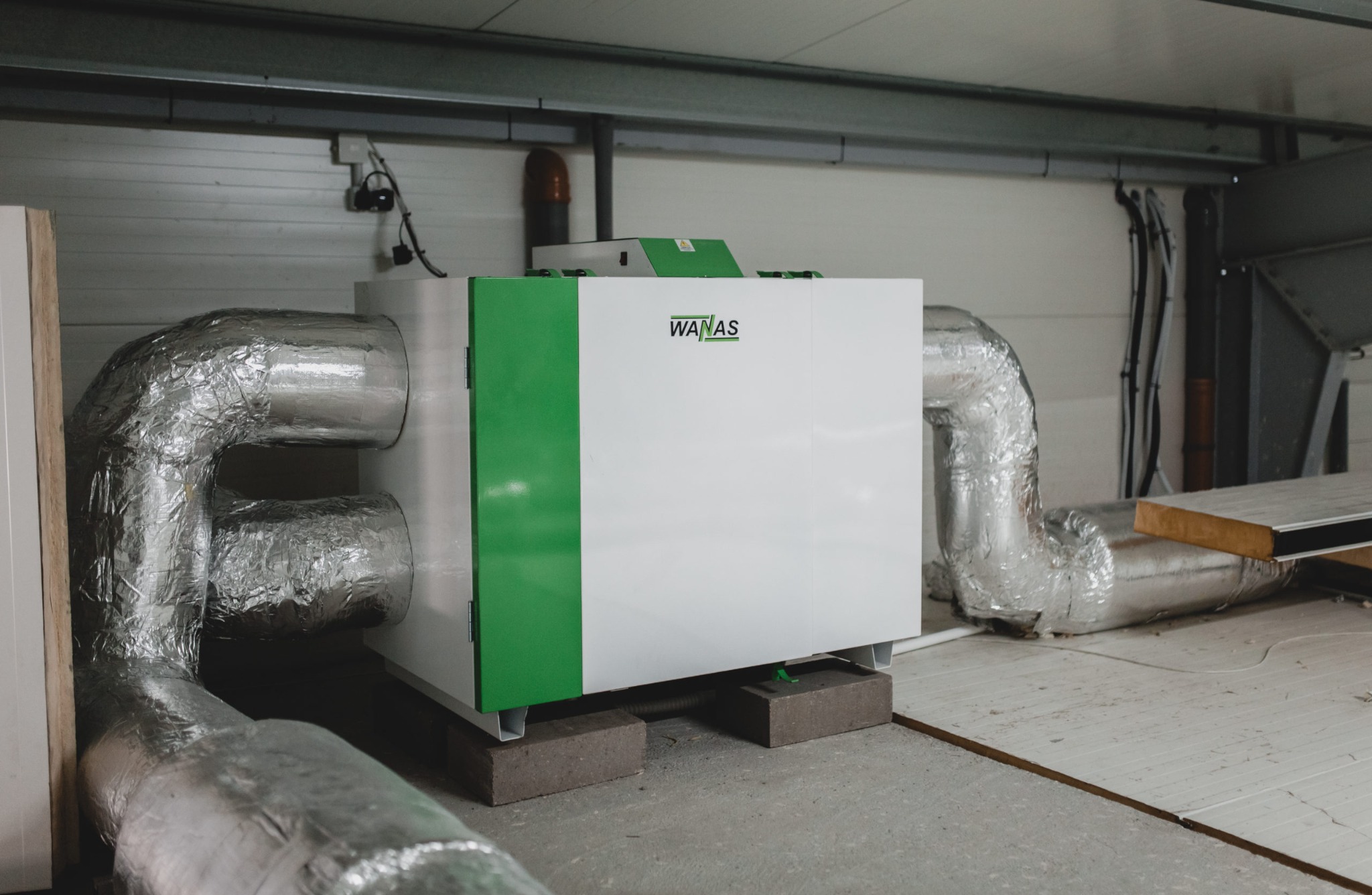 Centrala wentylacyjna WANAS z izolowanymi przewodami w pomieszczeniu technicznym, ustawiona na betonowych bloczkach.