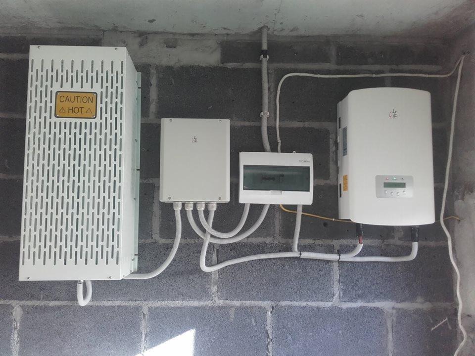 Elementy instalacji na szarej ścianie z betonu komórkowego: duża obudowa z wentylacją i ostrzeżeniem o wysokiej temperaturze, mniejsza obudowa, skrzynka rozdzielcza i falownik z wyświetlaczem.