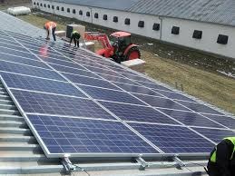 Montaż paneli słonecznych na dachu budynku gospodarczego, widoczne osoby w odblaskowych kamizelkach i czerwony traktor w tle.