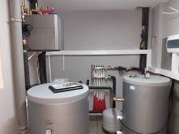 Zestaw grzewczy w kotłowni: szary piec, dwa srebrne zbiorniki akumulacyjne z manometrami, rozdzielacz ogrzewania podłogowego, rury i instalacja. Na jednym ze zbiorników leży tablet.