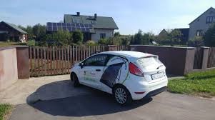 Biały samochód osobowy z grafiką paneli słonecznych na karoserii zaparkowany na podjeździe przed domem z instalacją solarną na dachu. W tle ogrodzenie i zabudowa jednorodzinna.