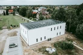 Biały budynek z panelami fotowoltaicznymi na dachu, widok z lotu ptaka, obok parking z zaparkowanym samochodem, w tle zabudowa miejska i drzewa.