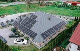 Dom jednorodzinny z szarym dachem pokrytym panelami słonecznymi, widok z góry, otoczony zielonym trawnikiem i tujami, zaparkowane samochody na podjeździe.