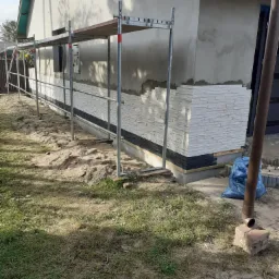 Montaż dekoracyjnej okładziny z białych i czarnych płytek imitujących kamień na elewacji domu jednorodzinnego, widoczne rusztowanie, przygotowany teren wokół budynku.
