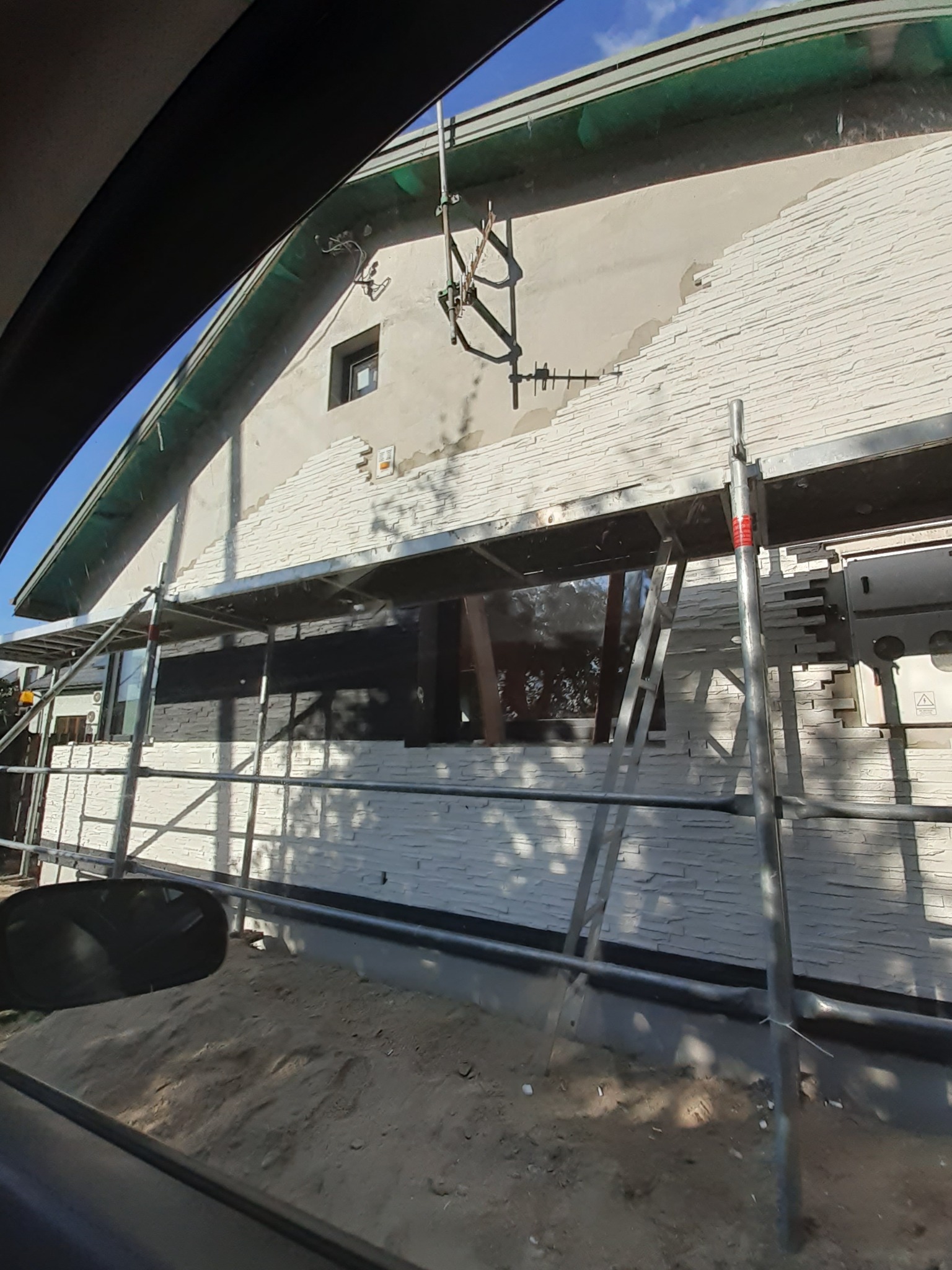 Fragment elewacji budynku w trakcie wykańczania z użyciem białej okładziny imitującej kamień, widoczne rusztowanie i fragment okna, ujęcie zza szyby samochodu.