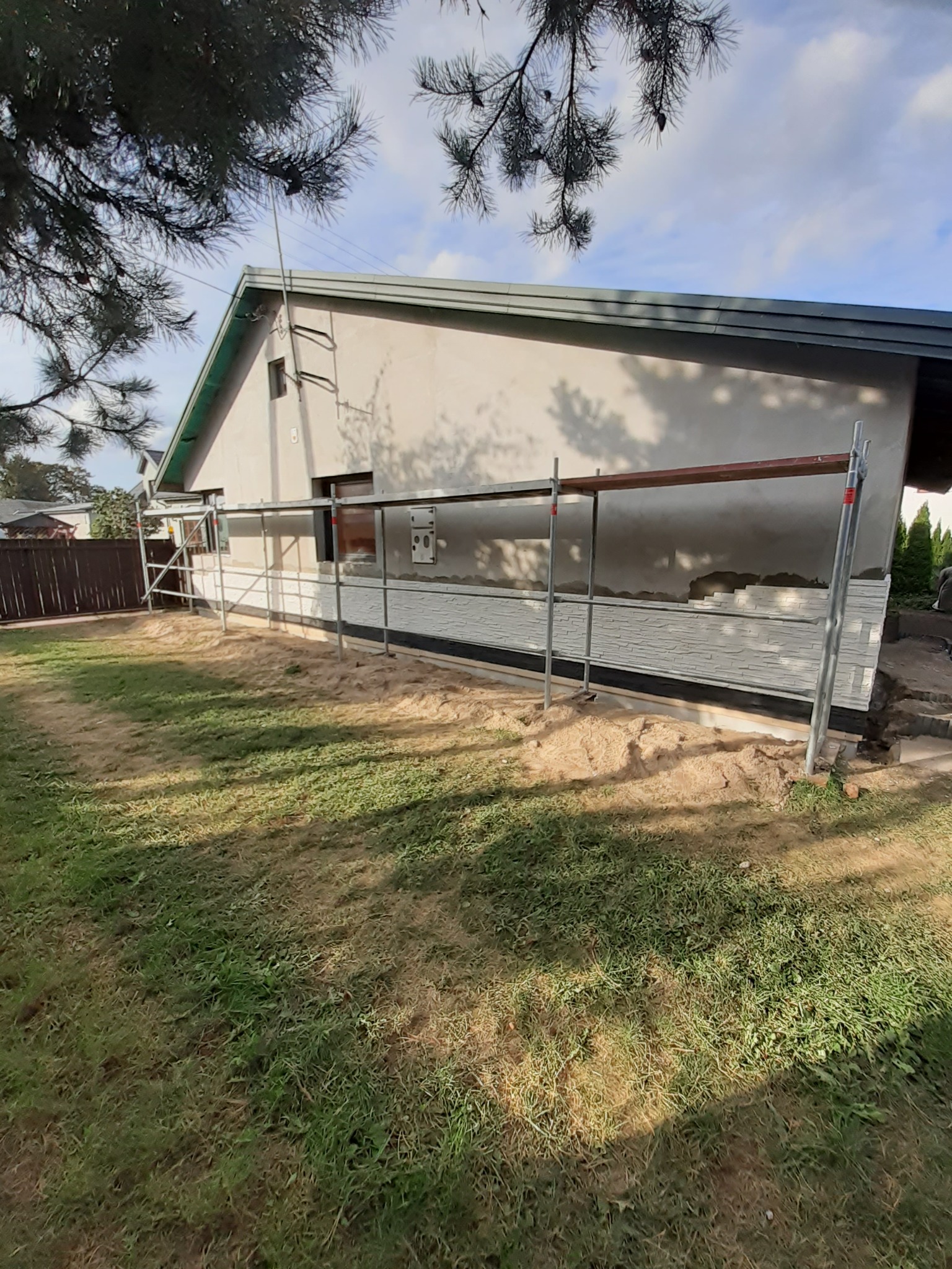 Dom jednorodzinny w trakcie wykańczania elewacji, widoczne rusztowania, fragment ściany z zamontowaną imitacją kamienia oraz ociepleniem, trawnik z piaskiem i fragmenty drzewa w tle.