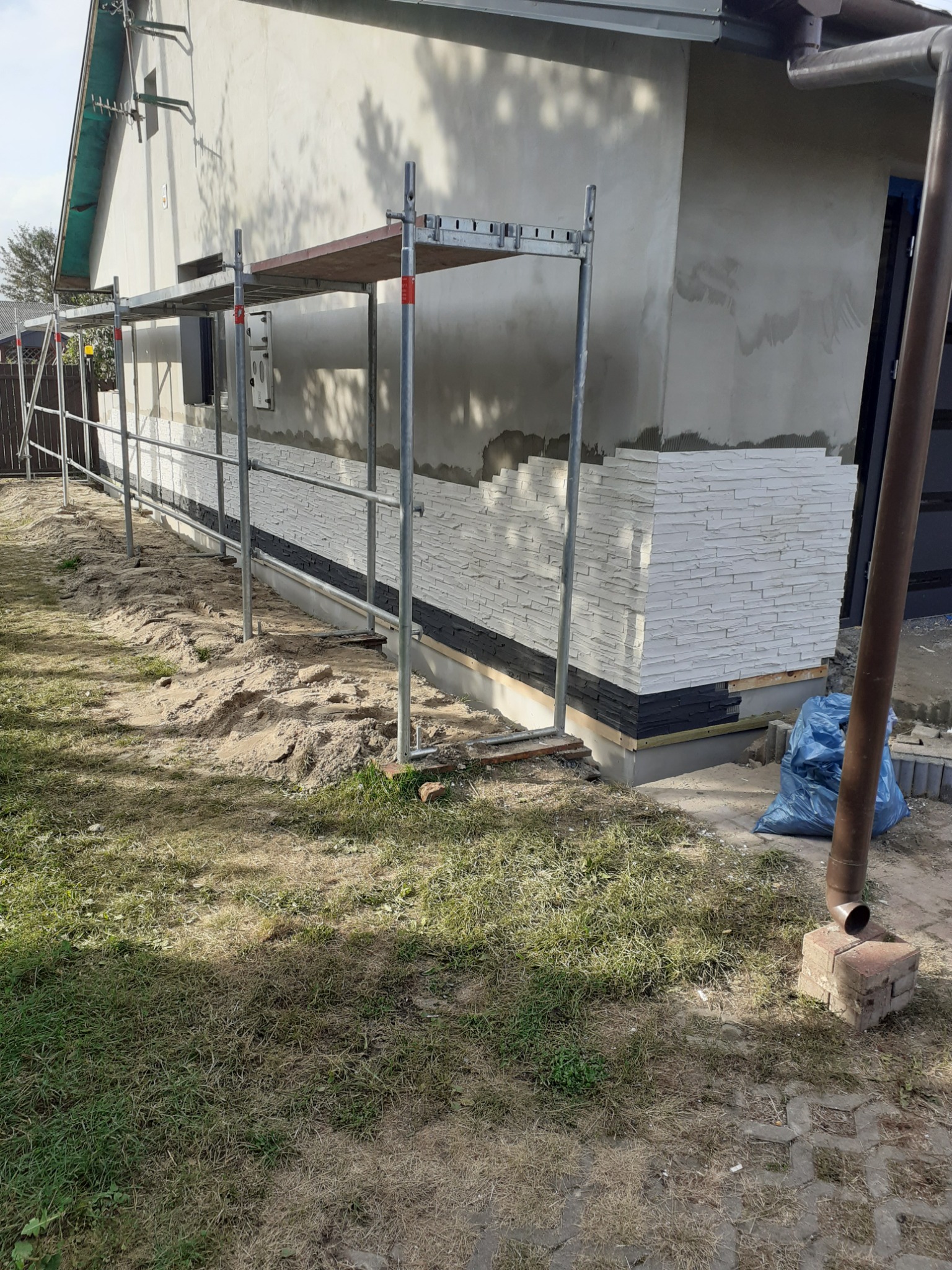 Montaż dekoracyjnej okładziny z białych i czarnych płytek imitujących kamień na elewacji domu jednorodzinnego, widoczne rusztowanie, przygotowany teren wokół budynku.