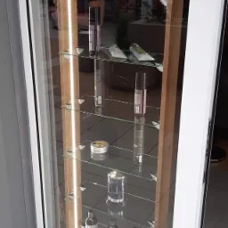 Wysoka, przeszklona witryna z drewnianą ramą i oświetleniem LED, prezentująca kosmetyki na szklanych półkach.