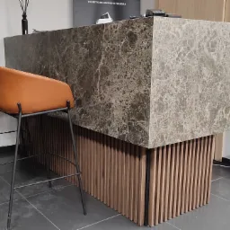 Recepcja z blatem z szarego marmuru, podświetlona listwami z jasnego drewna, z dwoma brązowymi krzesłami barowymi na metalowych nogach.