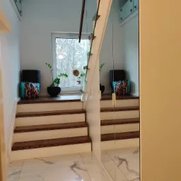 Nowoczesne schody drewniane z białymi podstopnicami i szklaną balustradą, z rośliną w doniczce i ozdobną torbą umieszczonymi na podeście okiennym, odbicie schodów i podestu w lustrzanej szafie.