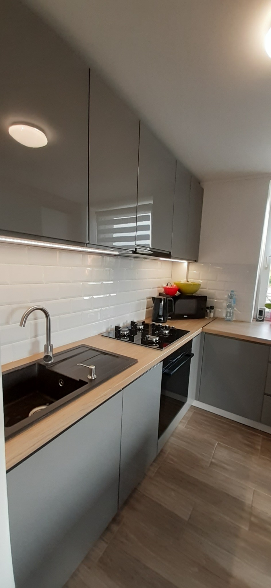 Szara kuchnia z drewnianym blatem, czarnym zlewem i płytą grzewczą, białe kafelki na ścianie, oświetlenie LED pod szafkami, widok na okno z roletą.
