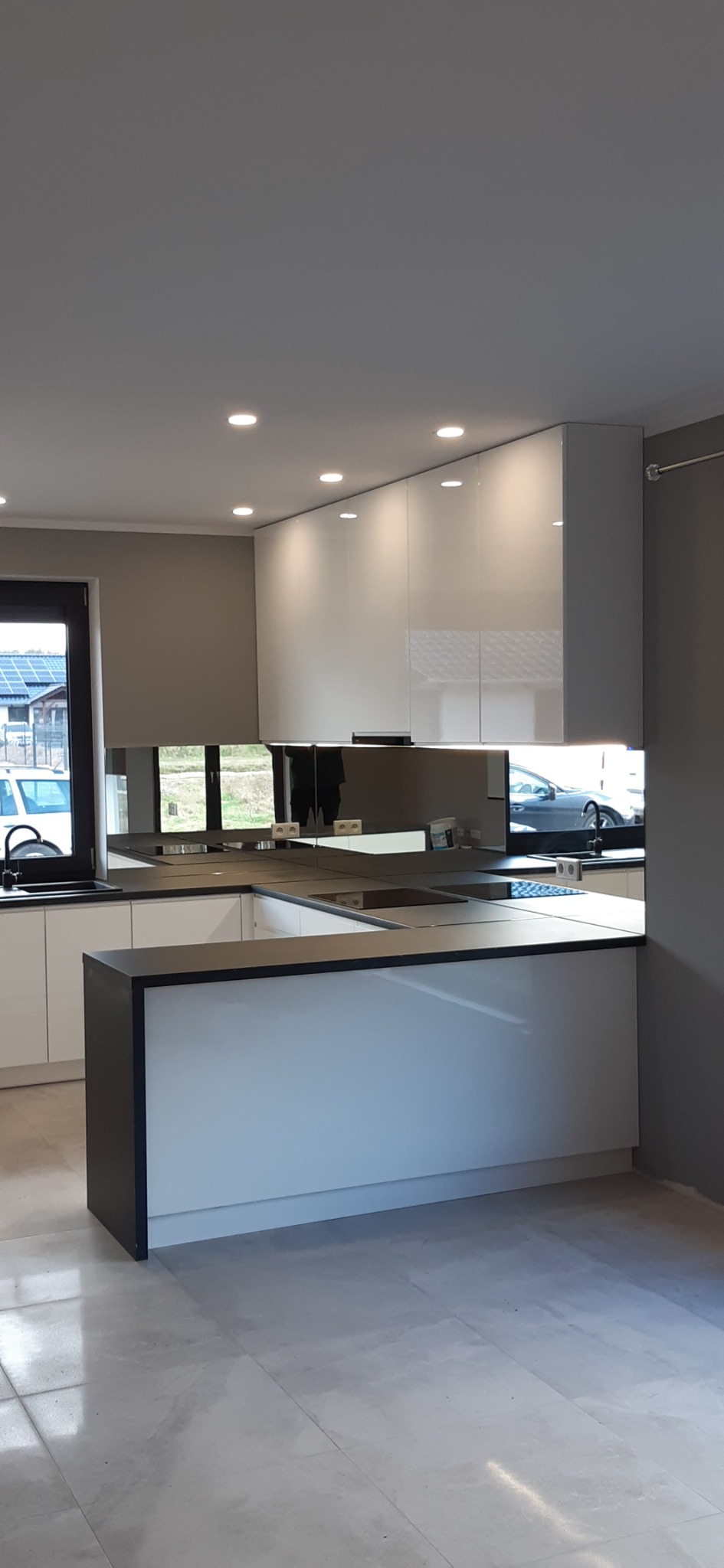 Nowoczesna kuchnia z białymi, lakierowanymi szafkami górnymi i wyspą z czarnym blatem, odbijająca się w lustrzanym panelu nad blatem roboczym, z widocznym fragmentem okna i samochodu za nim.